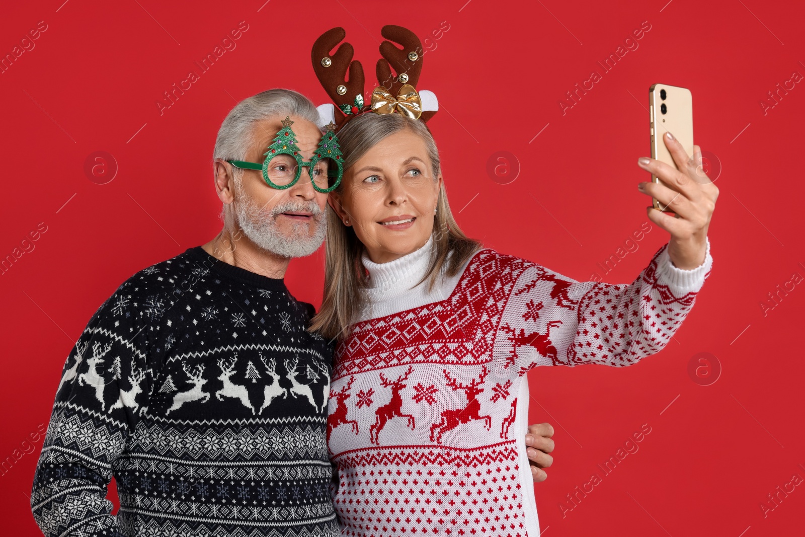 Photo of Senior couple in Christmas sweaters, reindeer headband and funny glasses taking selfie on red background