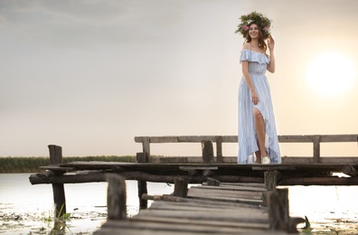 Photo of Young woman wearing wreath made of beautiful flowers on pier near river