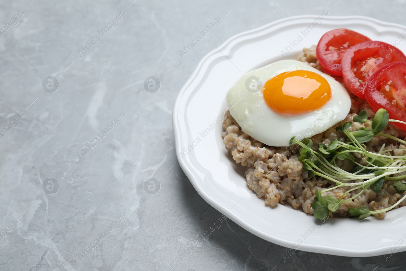 Photo of Delicious boiled oatmeal with fried egg, tomato and microgreens on light grey marble table, closeup. Space for text