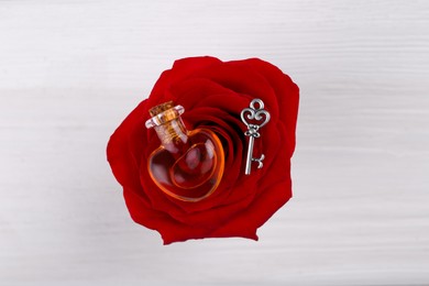 Photo of Heart shaped bottle of love potion with small key and red rose flower on white wooden table, top view