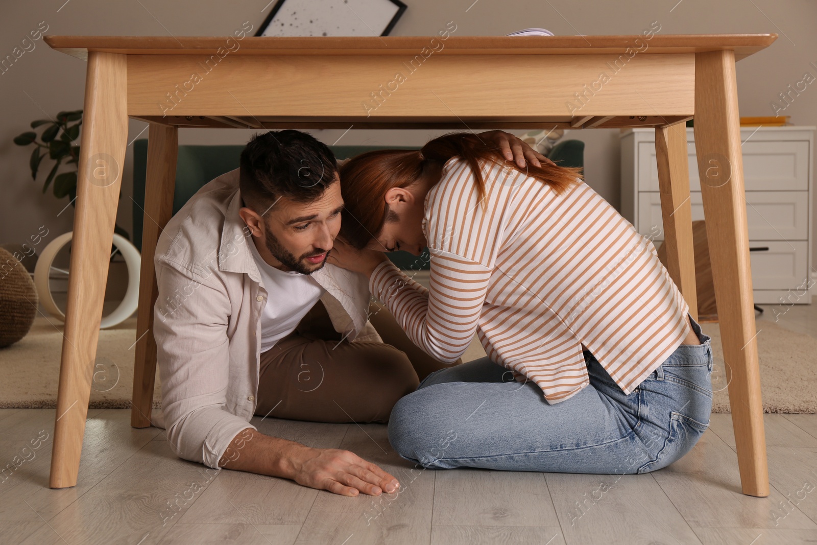 Photo of Scared couple hiding under table in living room during earthquake