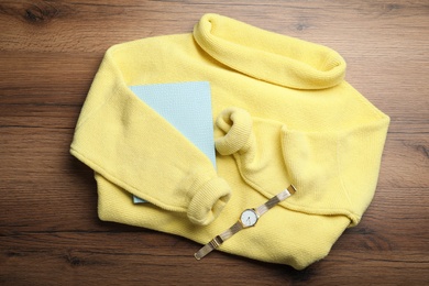 Yellow warm sweater with book and wristwatch on wooden table, flat lay