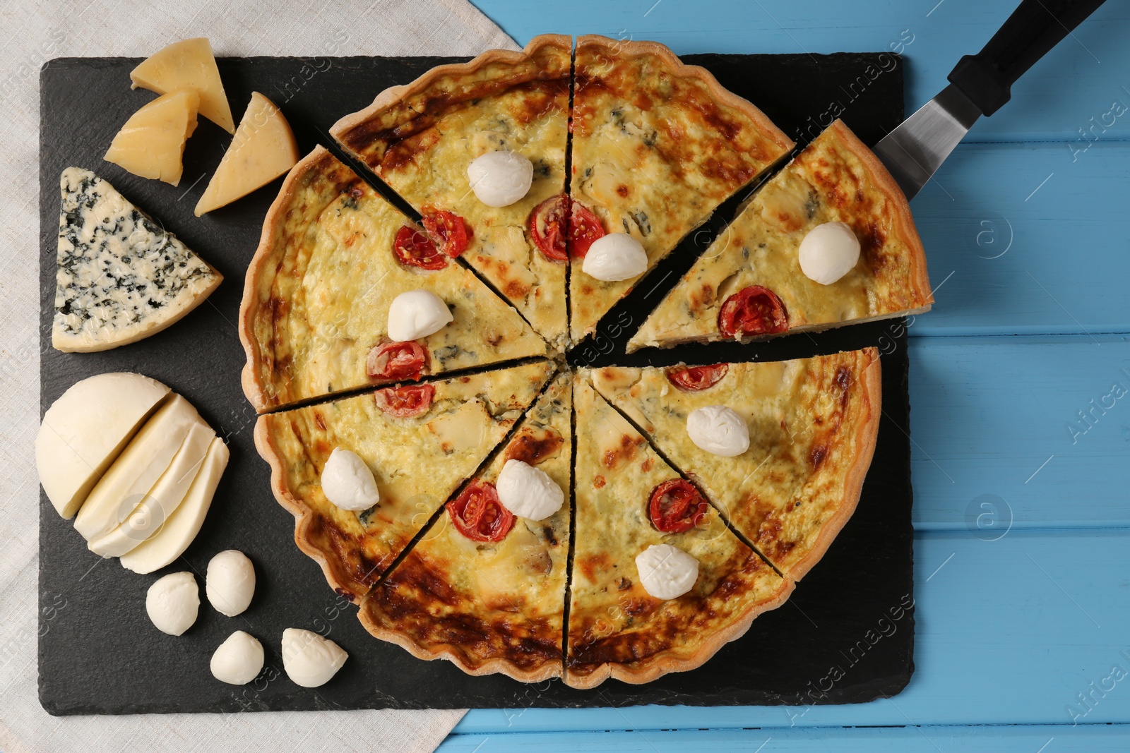 Photo of Taking piece of delicious homemade cheese quiche and ingredients on light blue wooden table, flat lay