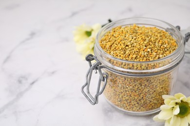 Fresh bee pollen granules in jar and flowers on light marble table, closeup. Space for text