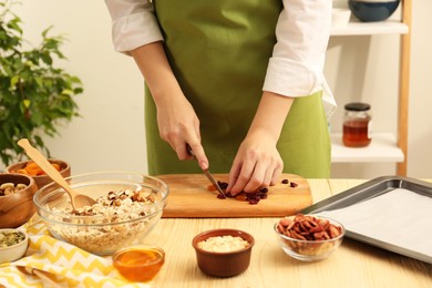 Photo of Making granola. Woman cutting dried apricots and cherries at table in kitchen, closeup