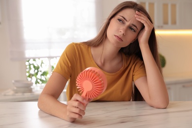 Photo of Woman with portable fan at table in kitchen. Summer heat