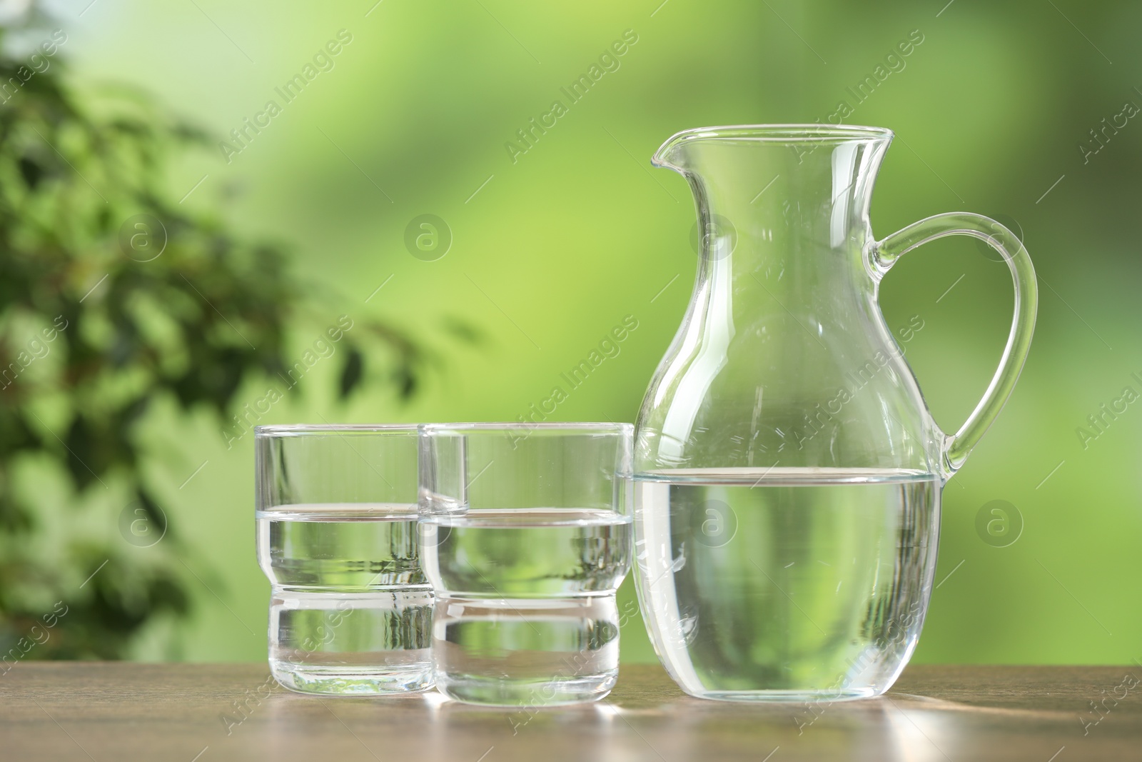 Photo of Jug and glasses with clear water on wooden table against blurred green background, closeup