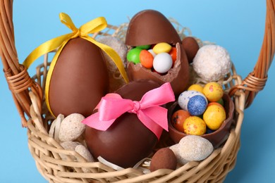 Photo of Wicker basket with tasty chocolate Easter eggs and different candies on light blue background, closeup