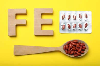 Photo of Wooden letters FE, spoon and pills on yellow background, flat lay. Anemia treatment