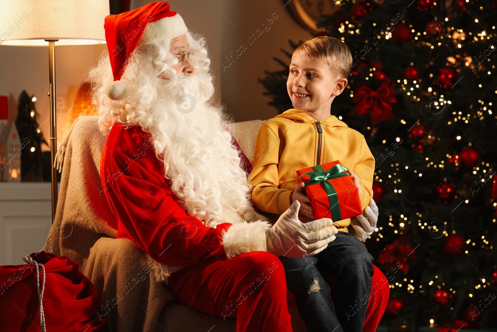 Photo of Merry Christmas. Smiling little boy sitting on Santa's knee and holding gift at home