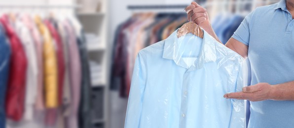 Dry-cleaning service. Man holding hanger with shirt in plastic bag indoors, space for text. Banner design