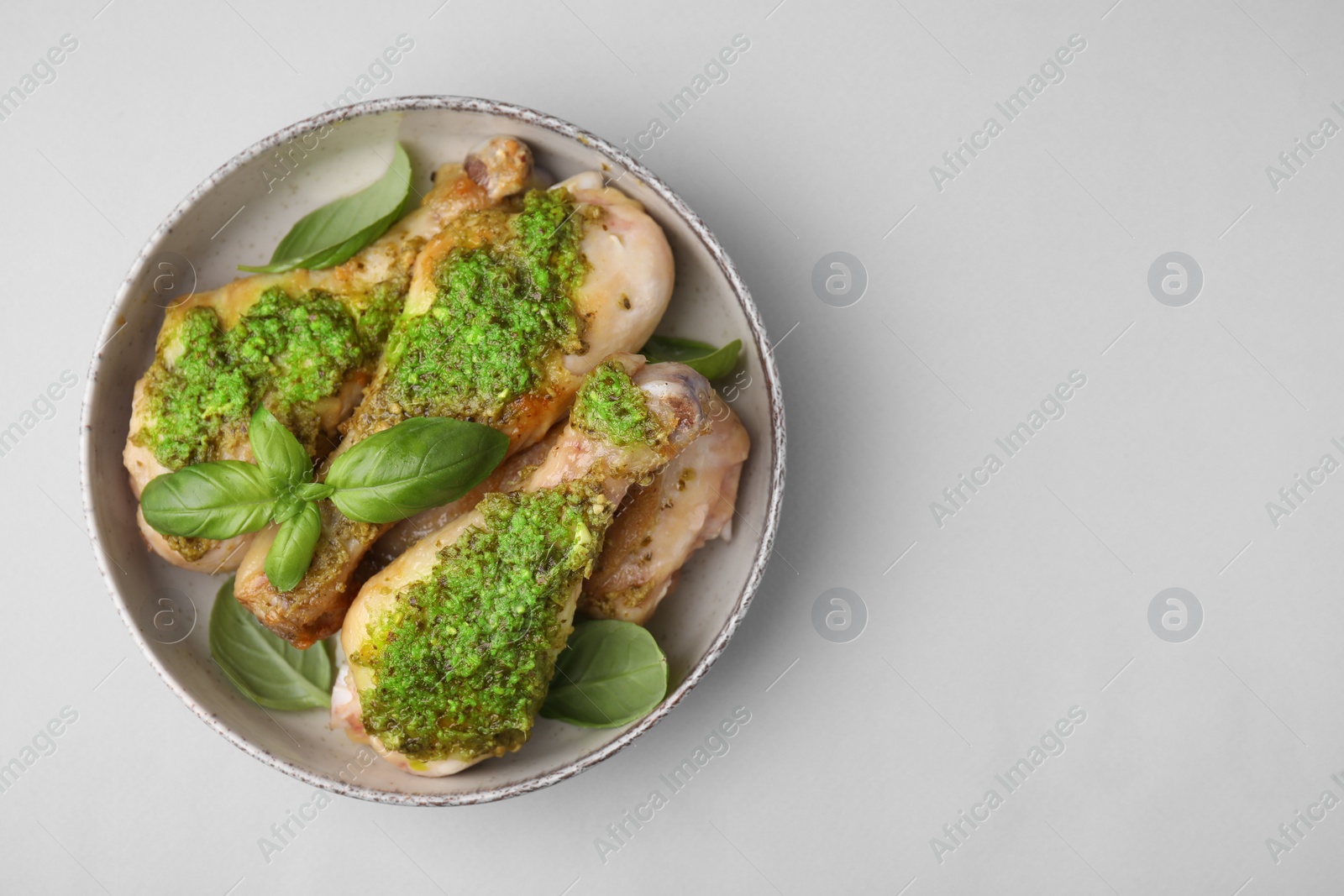 Photo of Delicious fried chicken drumsticks with pesto sauce and basil in bowl on light gray table, top view. Space for text