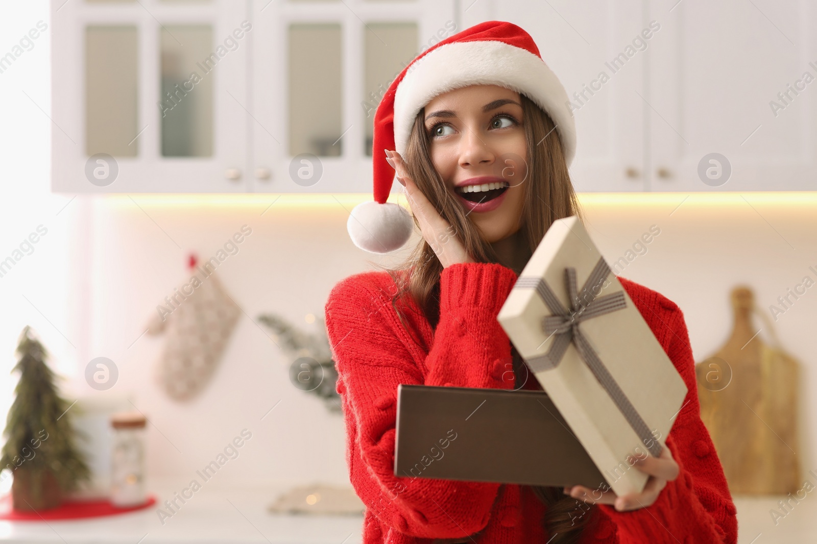 Photo of Emotional young woman in Santa hat opening present in kitchen, space for text. Celebrating Christmas