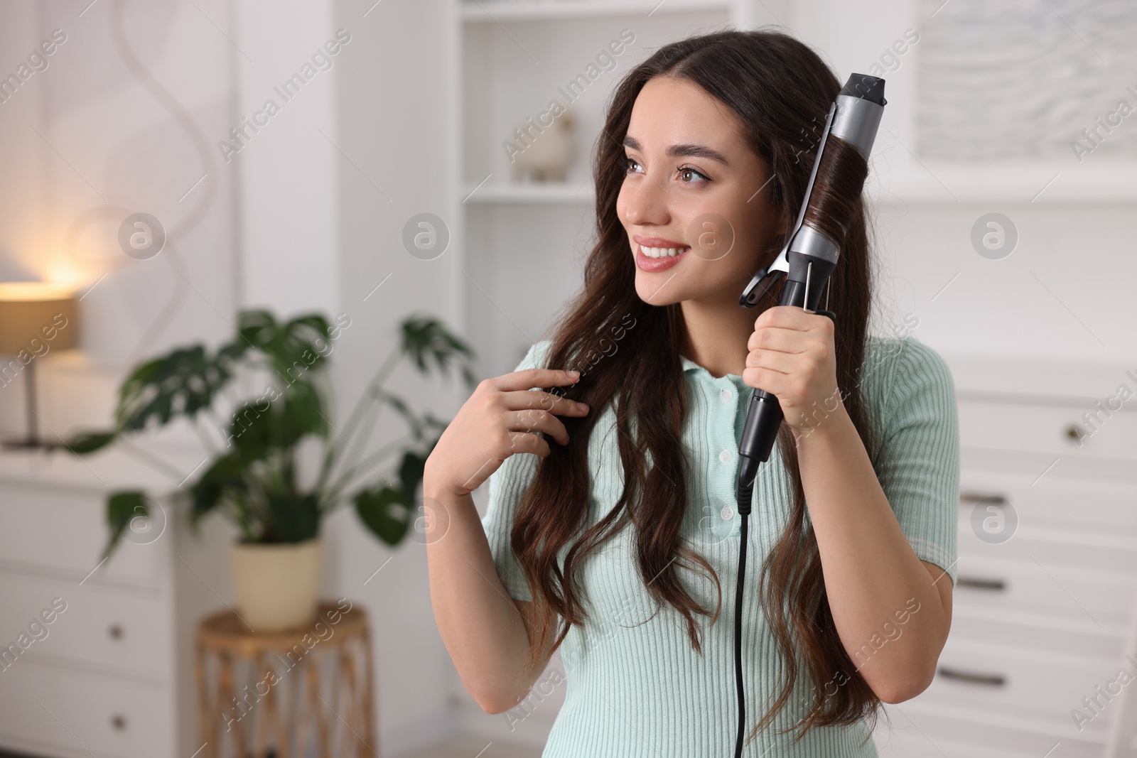 Photo of Smiling woman using curling hair iron at home. Space for text