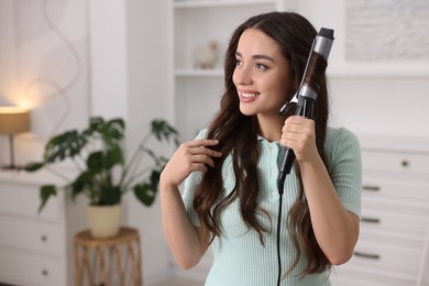 Smiling woman using curling hair iron at home. Space for text