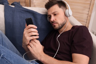 Upset man listening to music through headphones on sofa at home. Loneliness concept