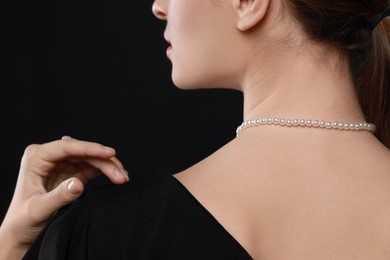 Young woman wearing elegant pearl necklace on black background, closeup
