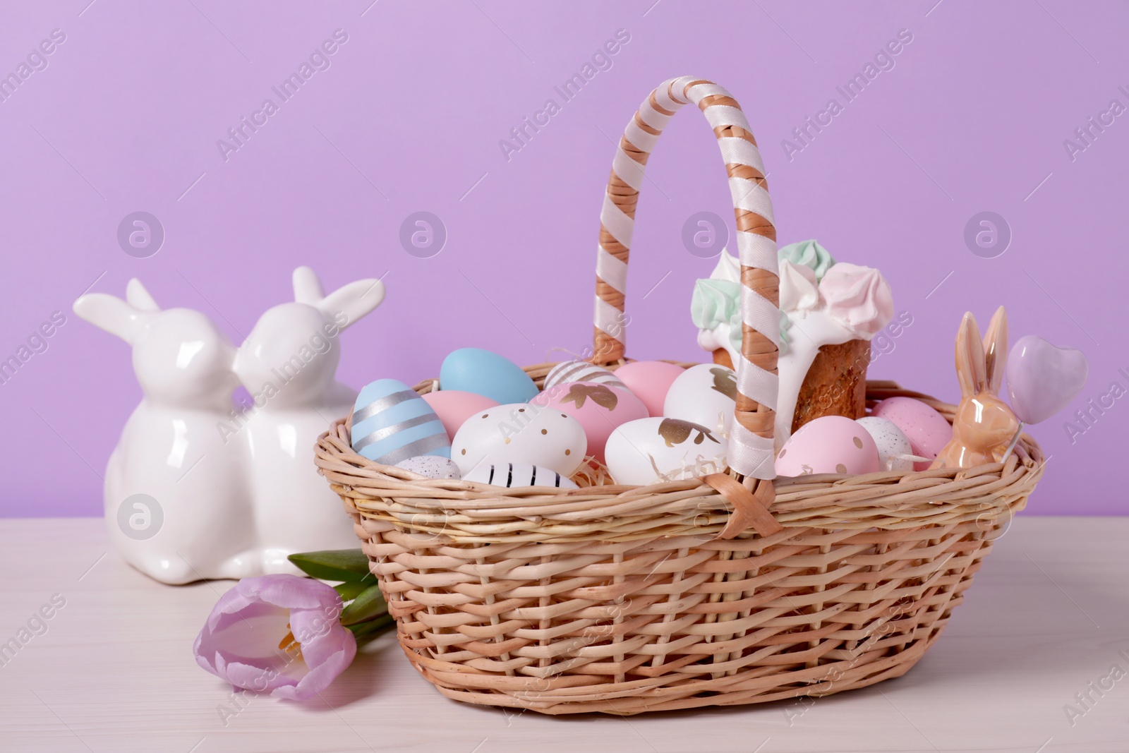 Photo of Easter basket with painted eggs, cake, flower and rabbits figure on white wooden table