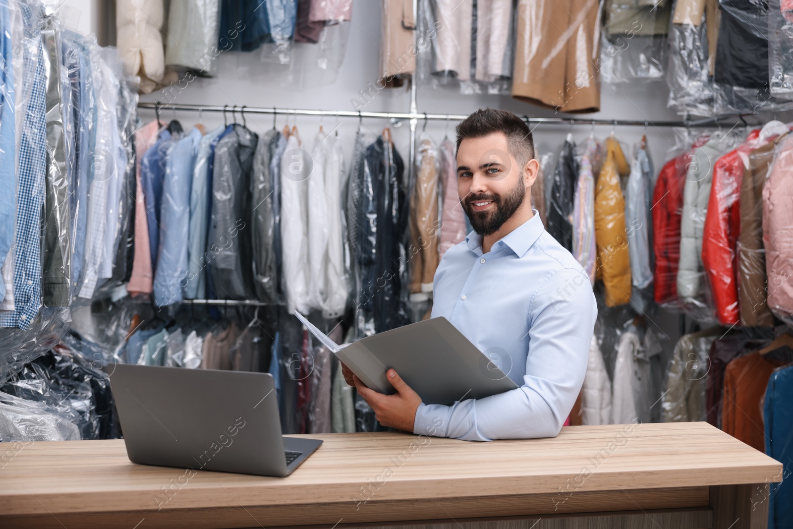 Photo of Dry-cleaning service. Happy worker with folder and laptop at counter indoors