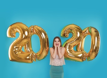 Photo of Excited young woman near golden 2020 balloons on turquoise background. New Year celebration