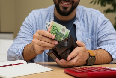 Young man putting money into wallet at table indoors, closeup