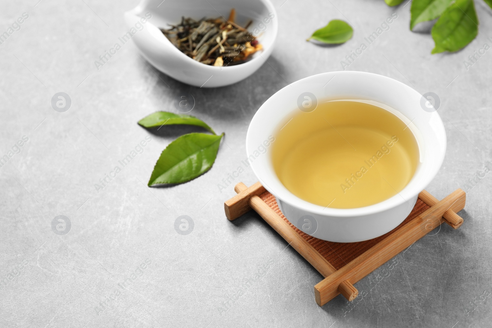 Photo of Cup of green tea, dry and fresh leaves on grey table. Space for text