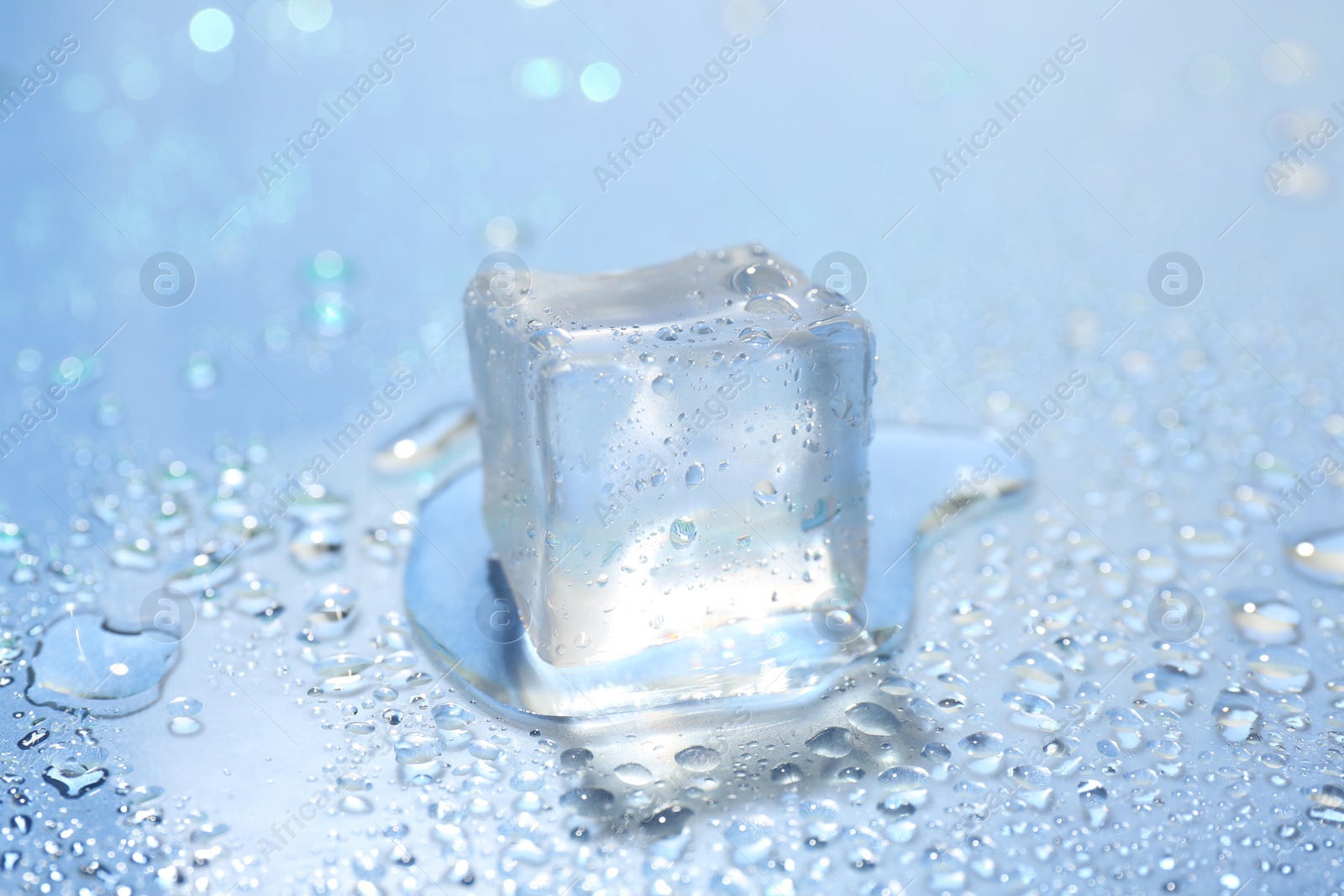 Photo of Melting ice cube and water drops on light blue background, closeup