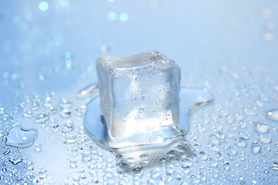 Photo of Melting ice cube and water drops on light blue background, closeup