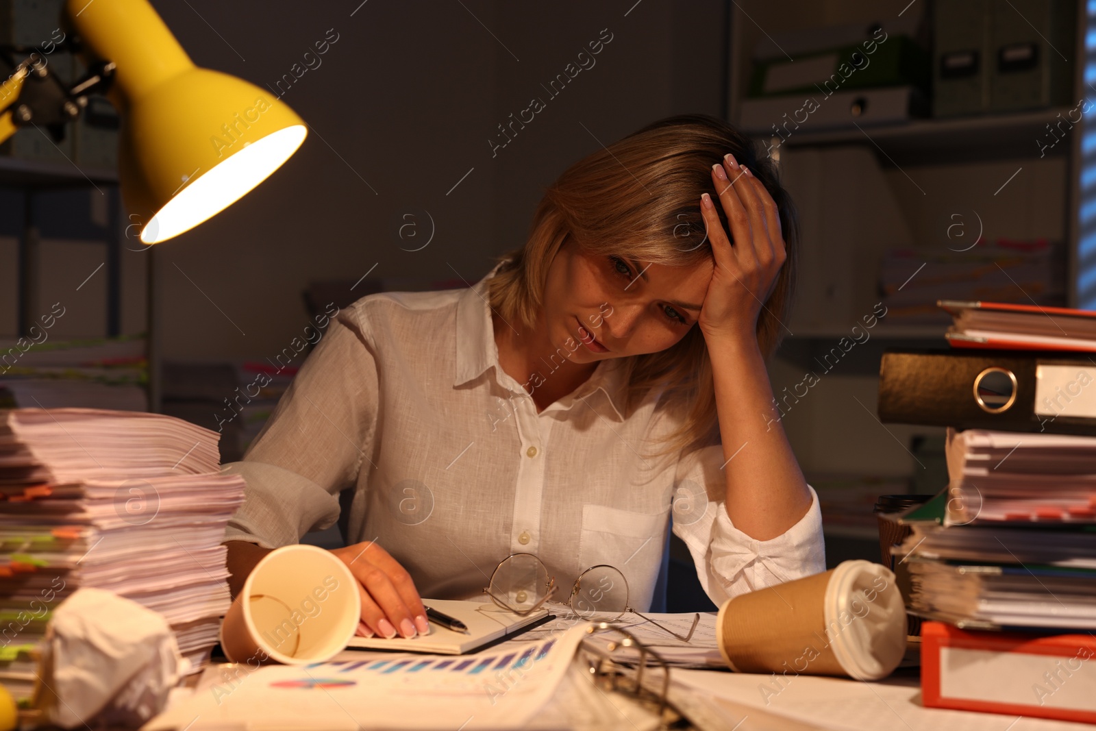 Photo of Overwhelmed woman surrounded by documents and paper coffee cups at table in office at night