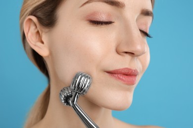 Young woman using metal face roller on light blue background, closeup