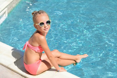 Photo of Happy cute little girl sitting at edge of swimming pool on sunny day. Space for text