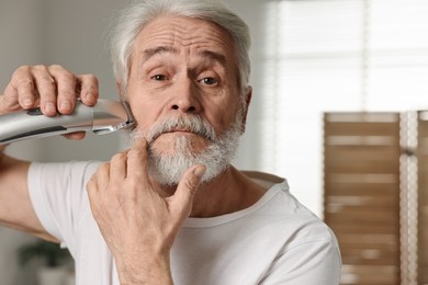 Photo of Senior man trimming beard with electric trimmer in bathroom