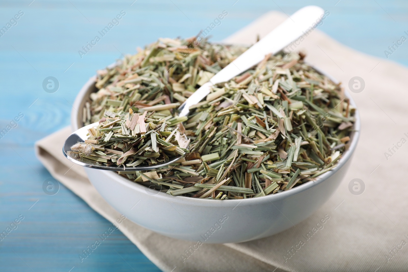 Photo of Bowl with aromatic dried lemongrass and spoon on light blue wooden table, closeup