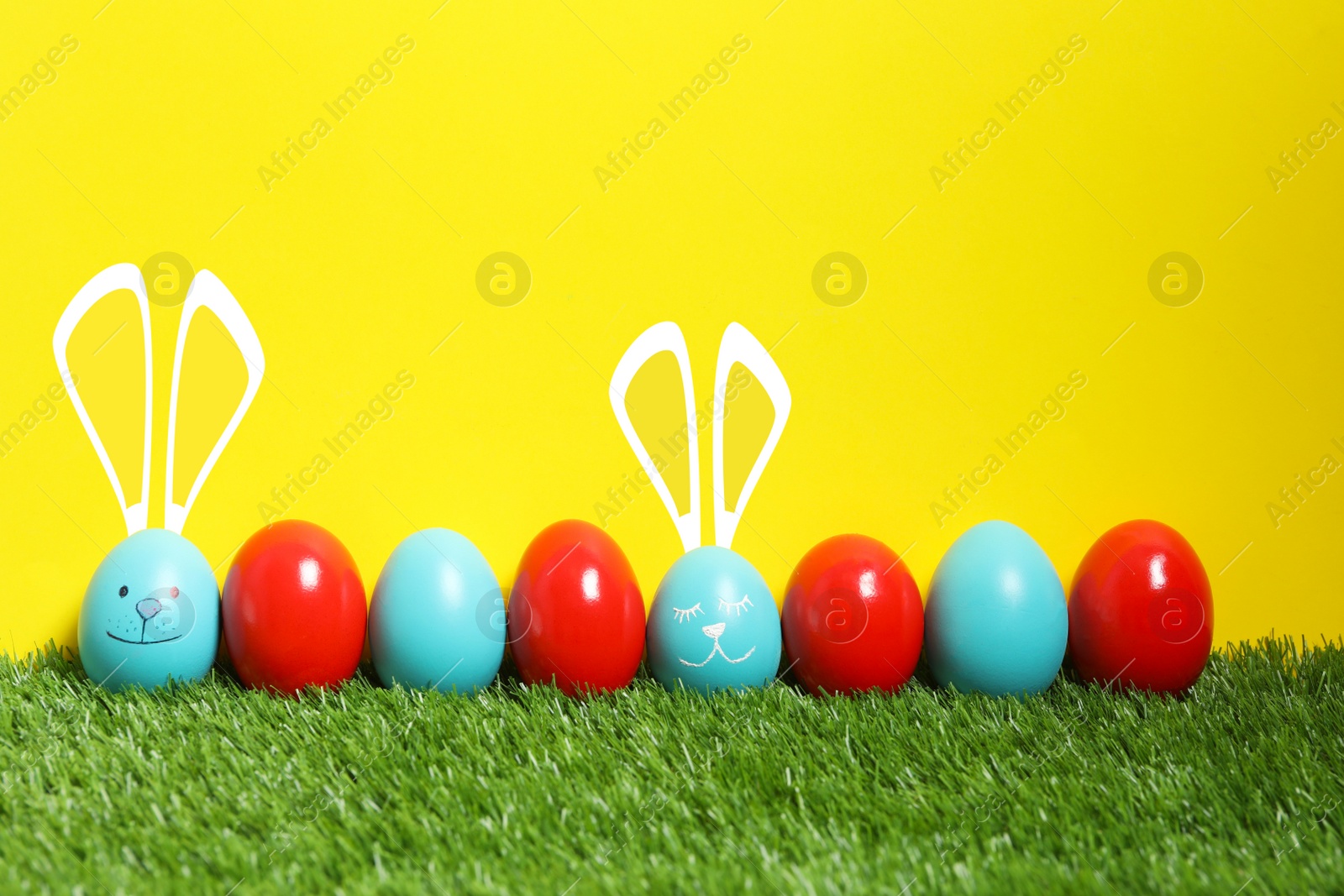 Image of Two eggs with drawn faces and ears as Easter bunnies among others on green grass against yellow background