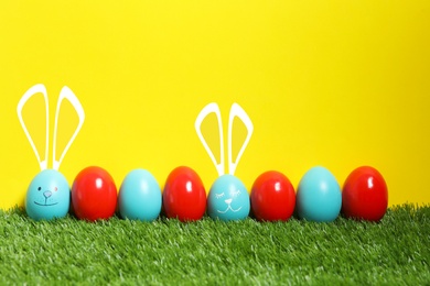 Image of Two eggs with drawn faces and ears as Easter bunnies among others on green grass against yellow background