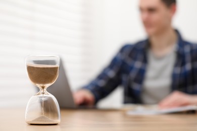 Hourglass with flowing sand on desk. Man taking notes while using laptop indoors, selective focus