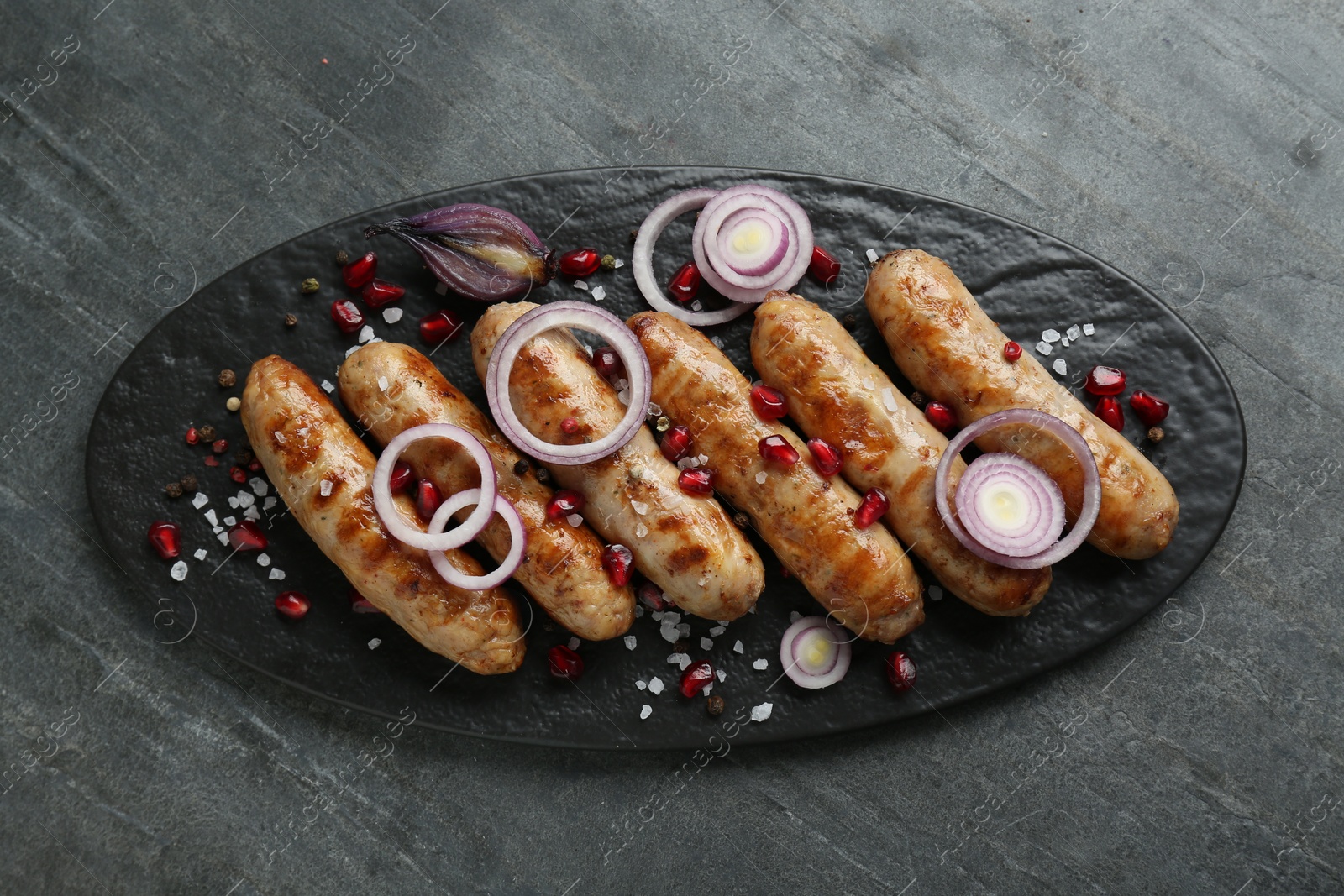 Photo of Tasty grilled sausages with onion rings and pomegranate seeds served on grey table, top view