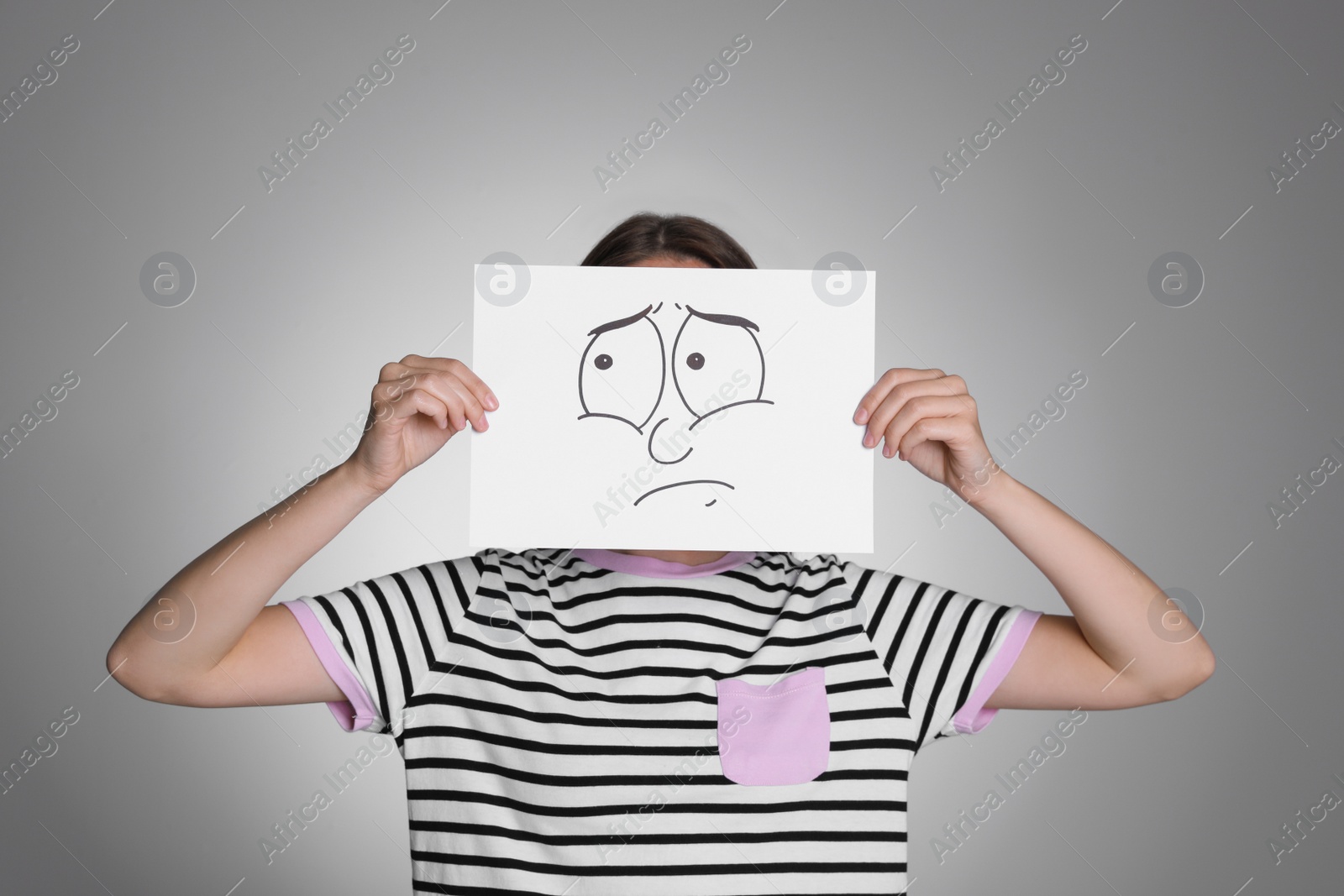 Photo of Woman hiding behind sheet of paper with sad face on grey background