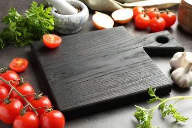 Photo of Black wooden cutting board, tomato, garlic and parsley on dark table, closeup