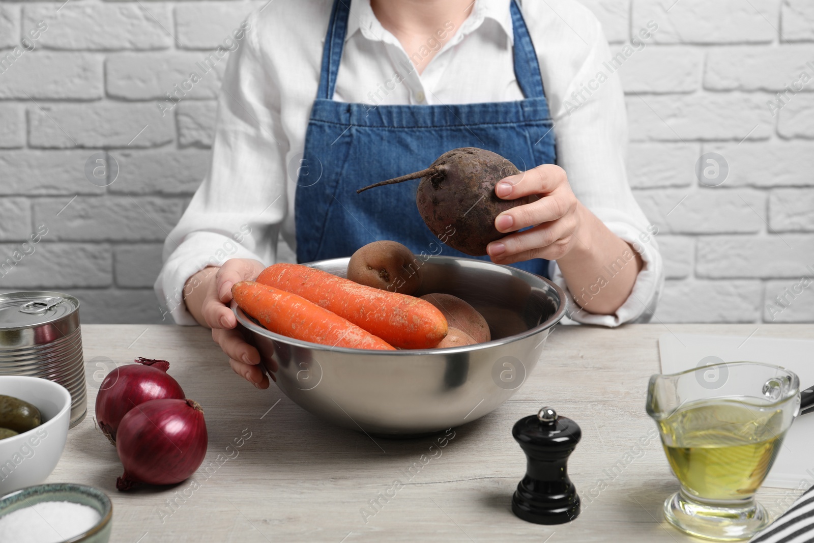 Photo of Woman putting beet into bowl with fresh vegetables at white wooden table, closeup. Cooking vinaigrette salad