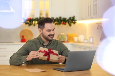 Photo of Celebrating Christmas online with exchanged by mail presents. Smiling man with gift box during video call on laptop at home