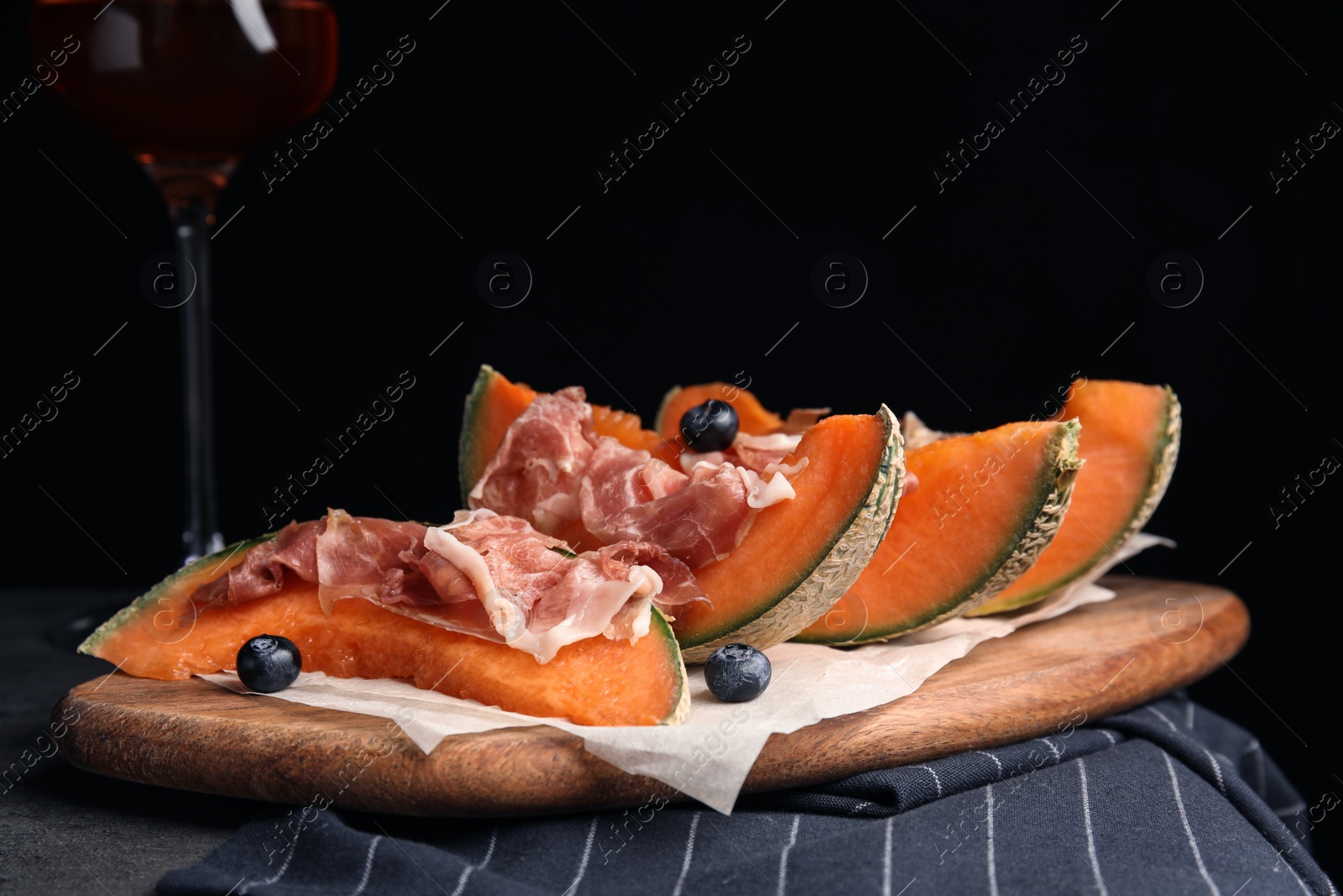 Photo of Wooden board with melon, prosciutto and blueberries on table against black background