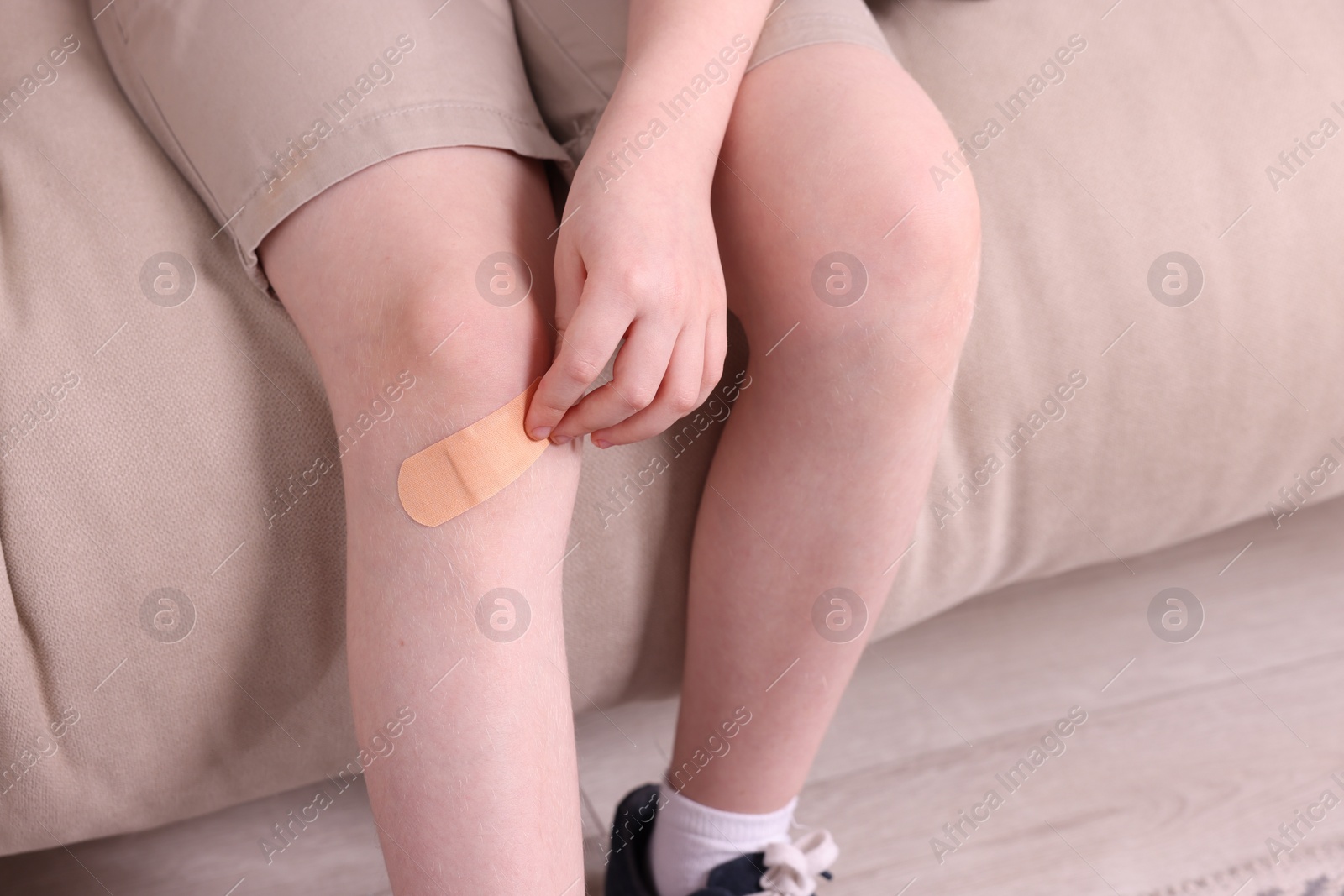 Photo of Little boy putting sticking plaster onto knee on sofa, closeup