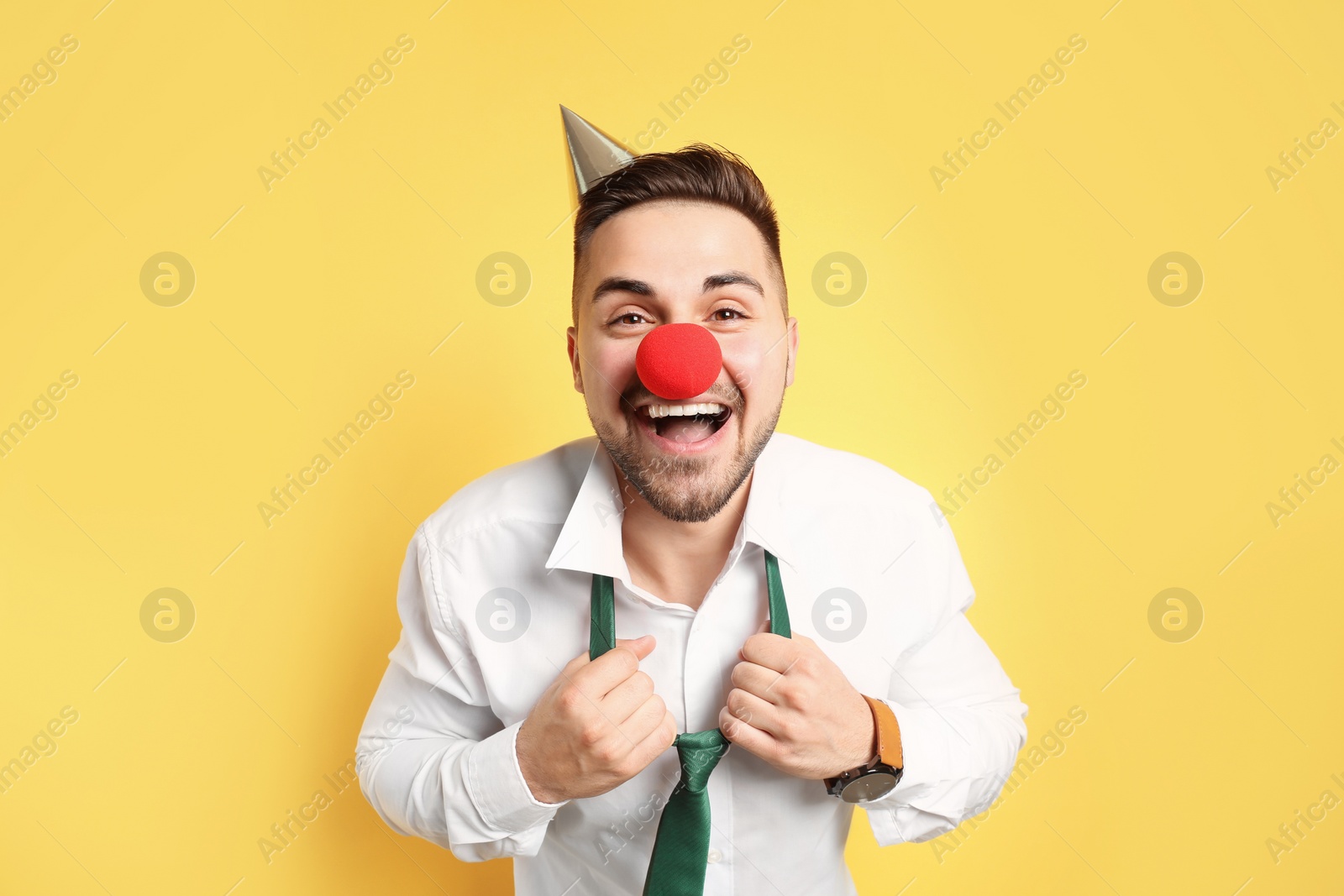 Photo of Emotional young man with party cap and clown nose on yellow background. April fool's day