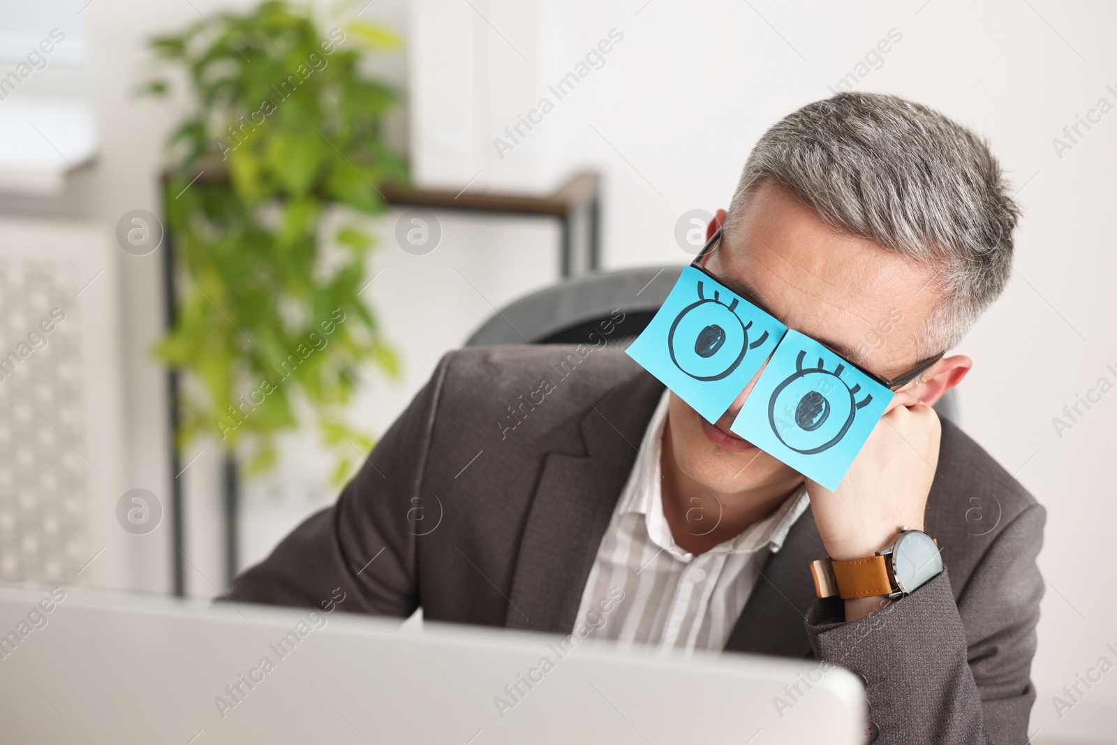 Photo of Man with fake eyes painted on sticky notes snoozing at workplace in office