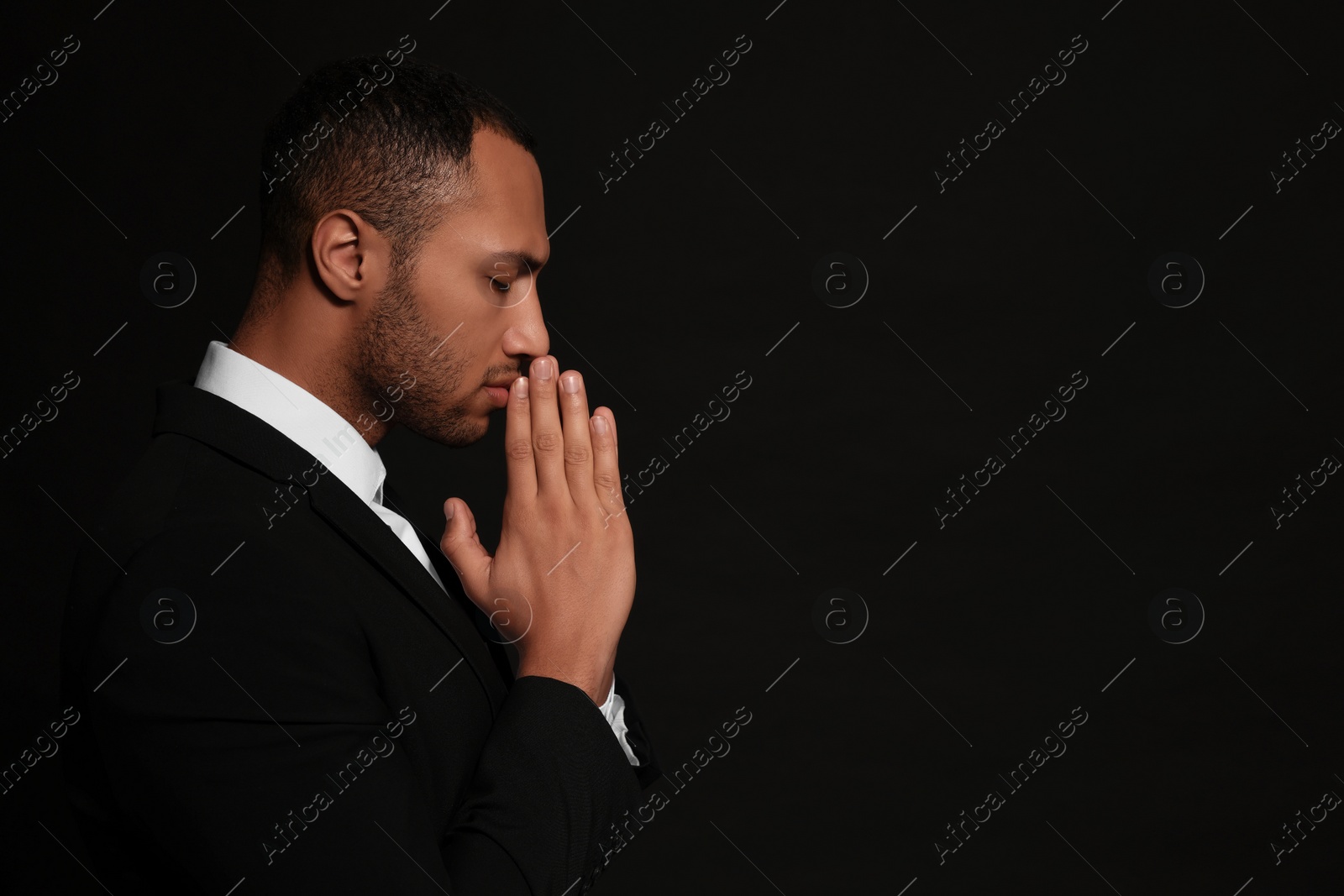 Photo of African American man with clasped hands praying to God on black background. Space for text