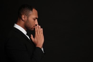 Photo of African American man with clasped hands praying to God on black background. Space for text