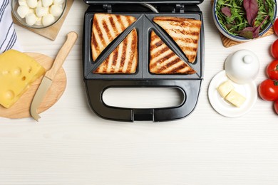 Modern sandwich maker with bread slices and different products on white wooden table, flat lay. Space for text