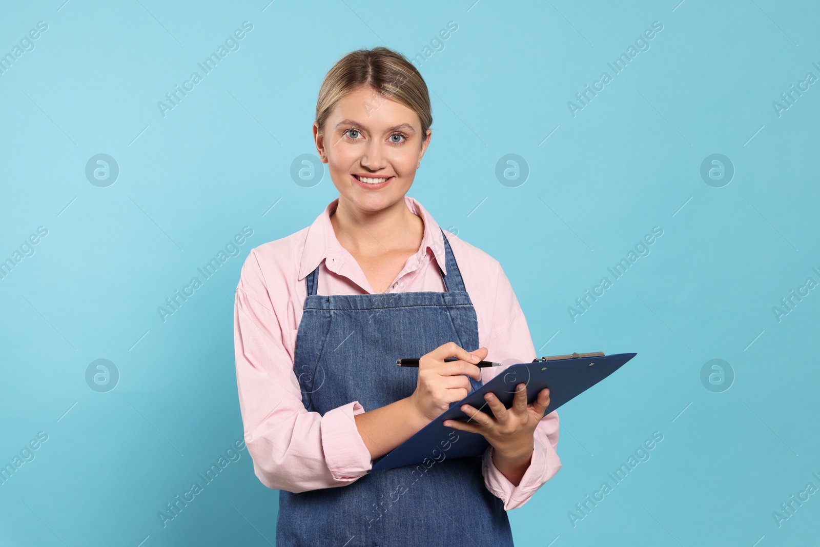 Photo of Beautiful young woman in denim apron with clipboard on light blue background