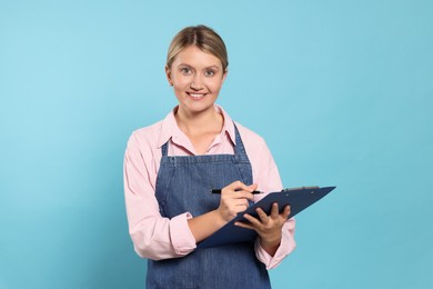 Photo of Beautiful young woman in denim apron with clipboard on light blue background